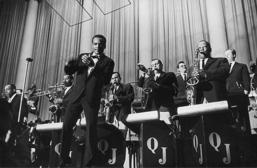 quincy jones jr stands on a stage in front of many musicians playing brass instruments