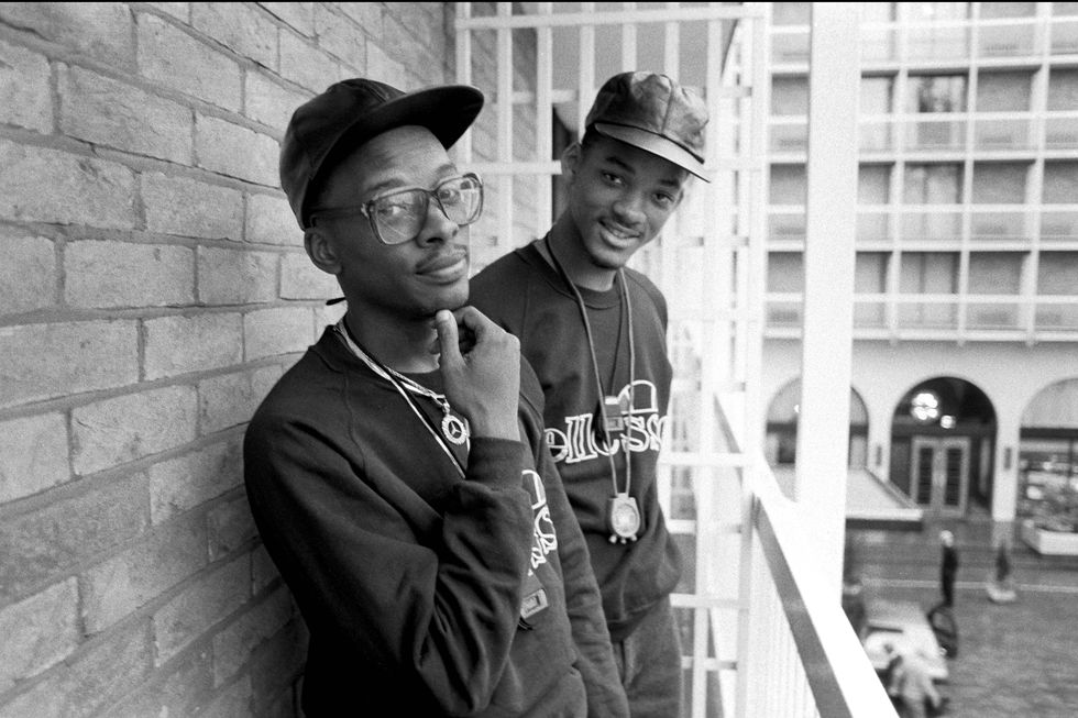 jeff townes aka dj jazzy jeff and will smith aka the fresh prince stand on a balcony against a brick wall and pose for a photo, townes holds his chin with one hand, smith smiles
