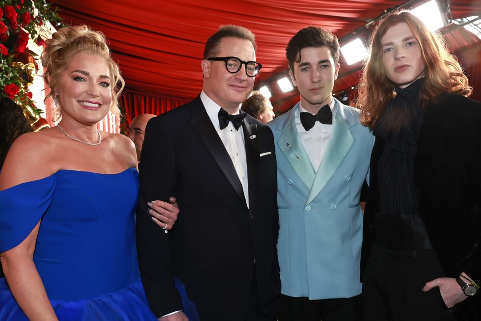 jeanne moore, brendan fraser, holden fraser, and leland fraser smile for a photo together while standing inside a red tent, moore wears a royal blue gown, brendan wears a black tuxedo, holden wears a baby blue suit jacket with a black bowtie and white shirt, leland wears an all black suit