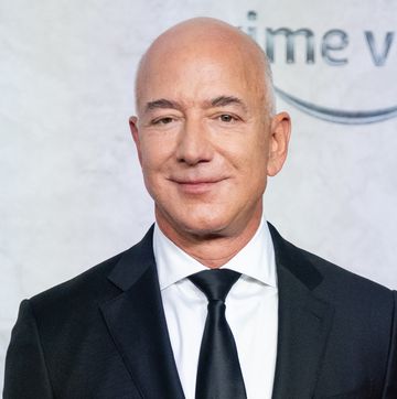 jeff bezos smiles at the camera, he wears a black suit and tie with a white collared shirt, behind him is a light gray background