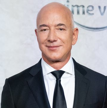 jeff bezos smiles at the camera, he wears a black suit and tie with a white collared shirt, behind him is a light gray background
