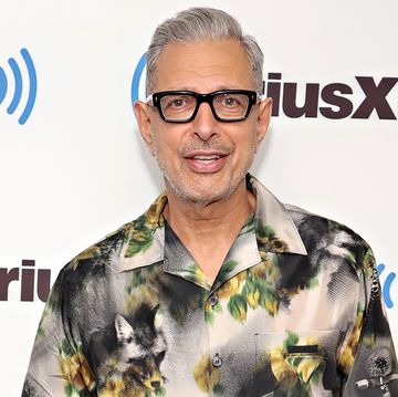 jeff goldblum, an older man stands looking at the camera with left hand in trouser pocket, greying hair, black thick rim glasses, wearing white and grey shirt with yellow flowers on and black trousers