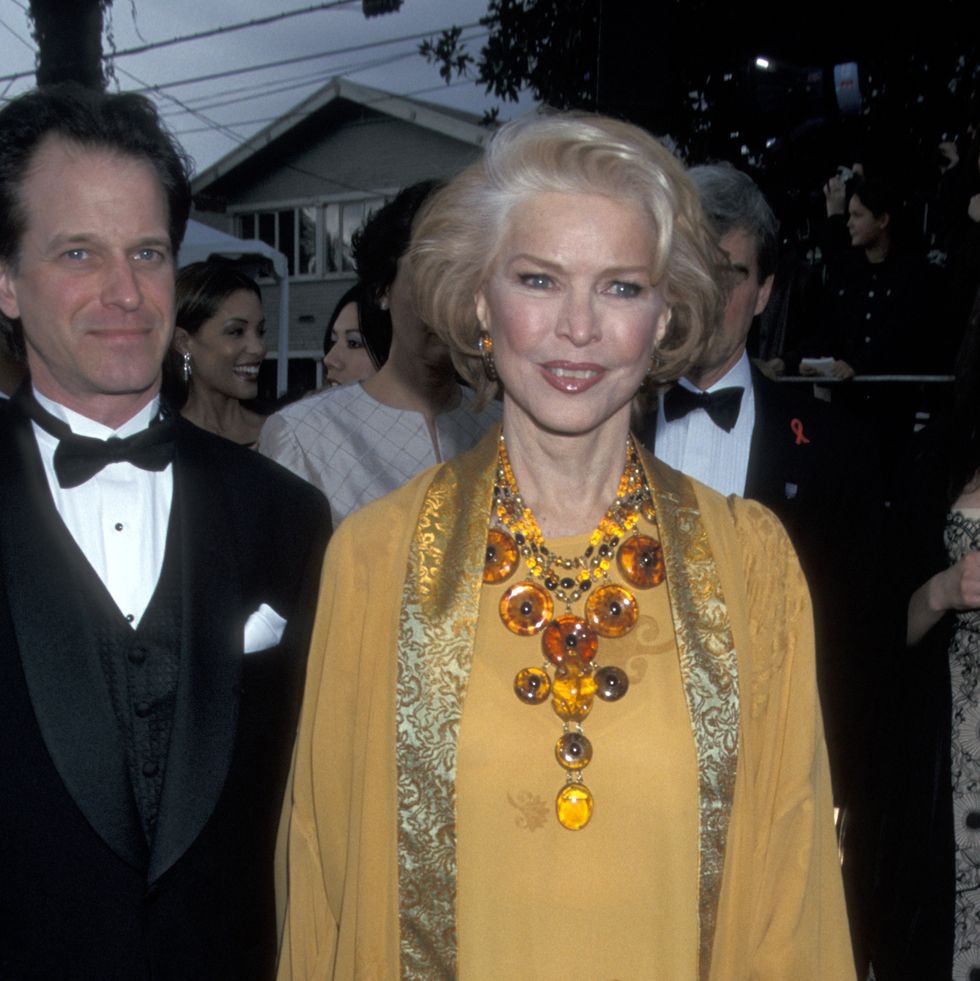 jefferson burstyn and ellen burstyn stand next to each other in front of a group of people, he wears a black tuxedo, she wears a yellow dress and caftan with a gold trim and a large yellow necklace