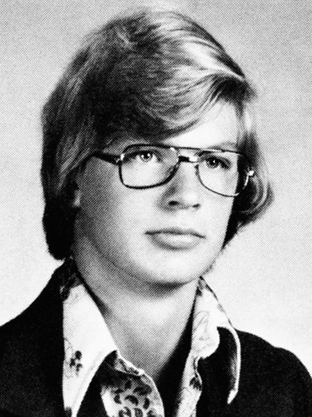 jeffrey dahmer poses for a black and white high school yearbook photo, he is looking off camera with a neutral expression on his face, he is wearing aviator glasses, a dark blazer, and a patterned shirt with a tall collar