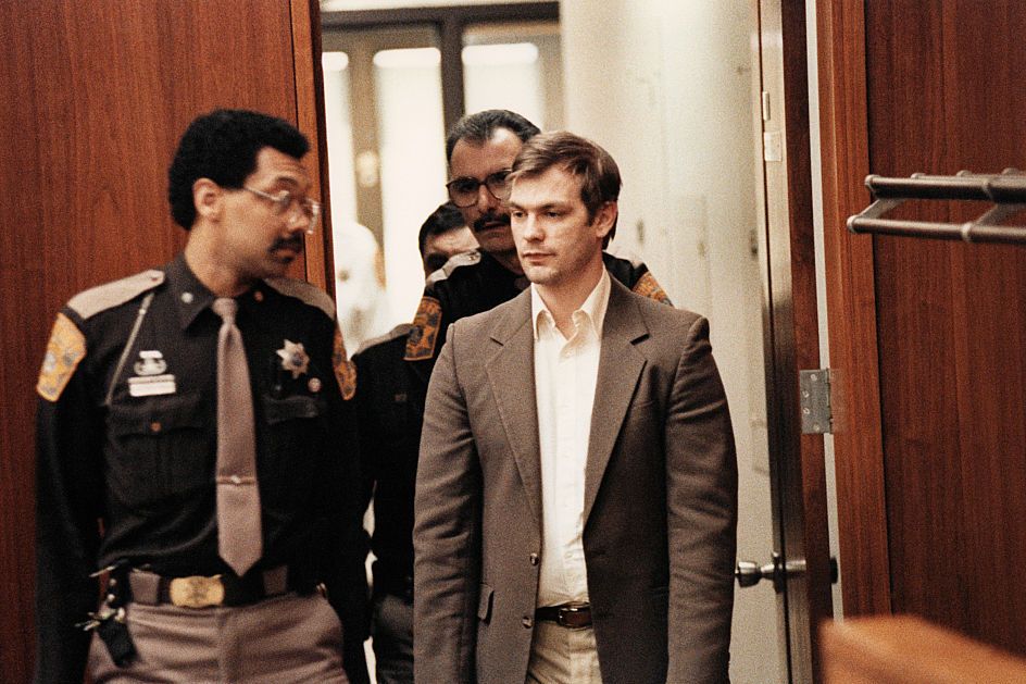 jeffrey dahmer, wearing a tan suit jacket and white shirt, is escorted by three uniformed police officers into a courtroom