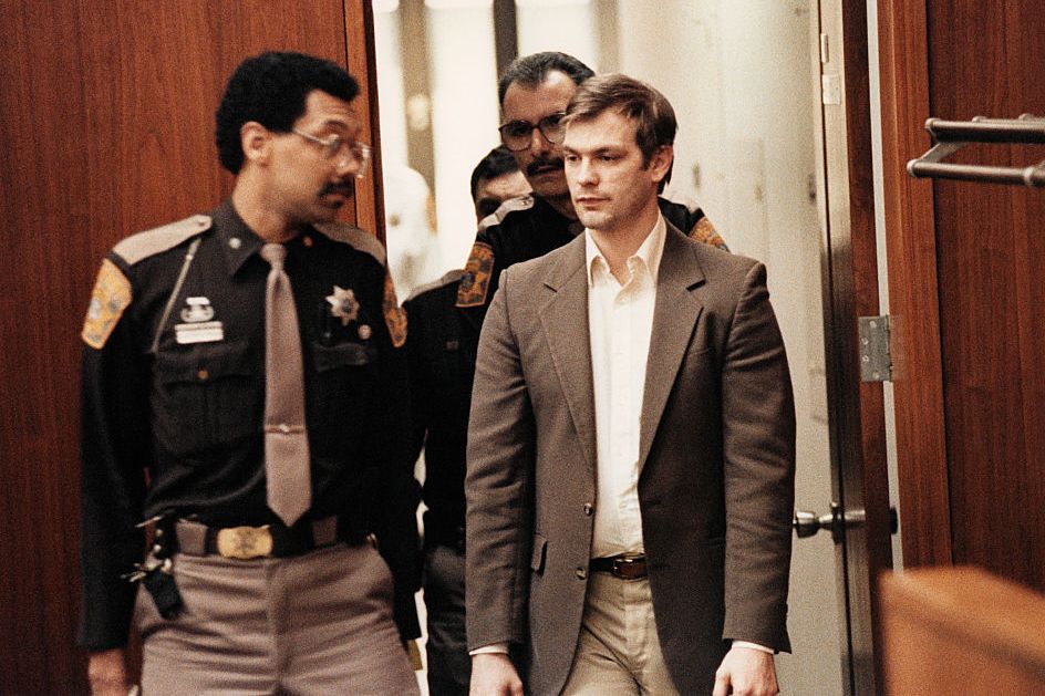 jeffrey dahmer, wearing a tan suit jacket and white shirt, is escorted by three uniformed police officers into a courtroom