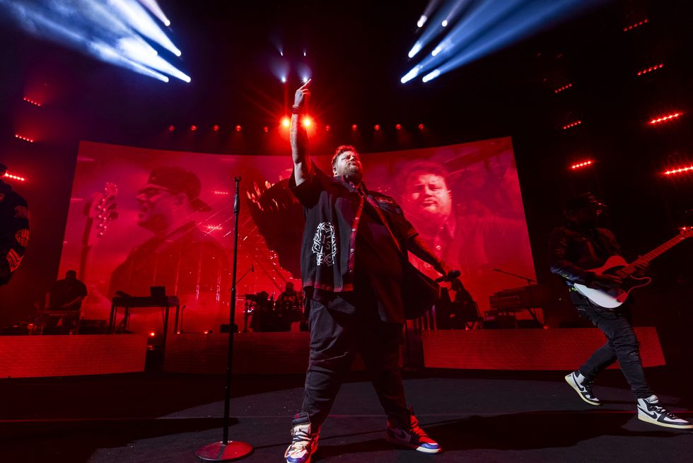 jelly roll stands on a stage with one hand pointing up in the air and another holding a microphone out at his side, he wears a black button up over a black shirt, dark pants, and sneakers, behind him is a large video screen and musicians with instruments