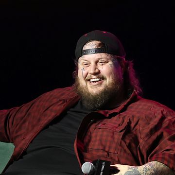 jelly roll sits on a green couch and smiles out to an unseen audience, he holds a microphone in one hand and wears a black outfit with a red plaid shirt unbuttoned and a black backward baseball hat