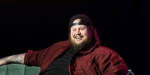 jelly roll sits on a green couch and smiles out to an unseen audience, he holds a microphone in one hand and wears a black outfit with a red plaid shirt unbuttoned and a black backward baseball hat