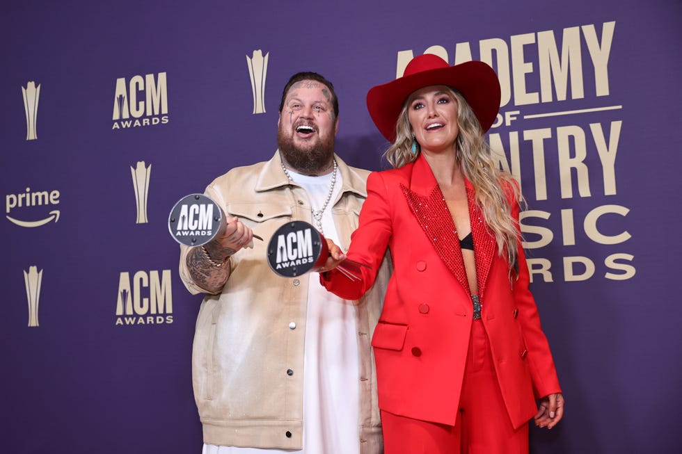 jelly roll and lainey wilson both hold up a silver trophy with one hand and smile while posing for photos in front of a purple backdrop