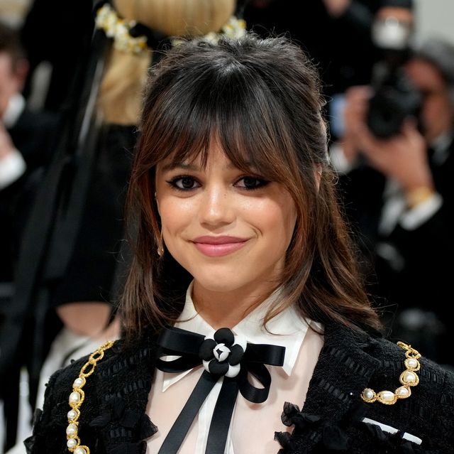 jenna ortega smiles at the camera, she wears a black and white formal outfit with a black bow in her white collared shirt, she has shoulder length brown hair and bangs