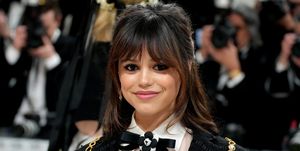 jenna ortega smiles at the camera, she wears a black and white formal outfit with a black bow in her white collared shirt, she has shoulder length brown hair and bangs