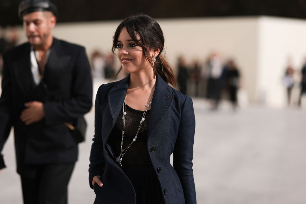 jenna ortega smiles and looks left of the camera, she stands outside with one hand in her jacket pocket, she also wears a black top and silver long necklace
