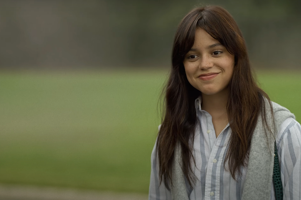 Jenna Ortega, eine Frau mit langen braunen Haaren, die ein weiß-blau gestreiftes Button-Down-Shirt trägt und mit Gras im Hintergrund lächelt, in einer Szene aus „Miller's Girl“.
