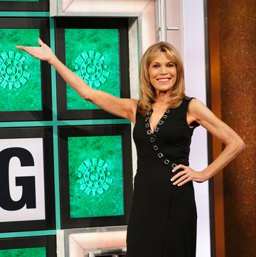 vanna white, wearing a black dress, gesturing toward the wheel of fortune puzzle