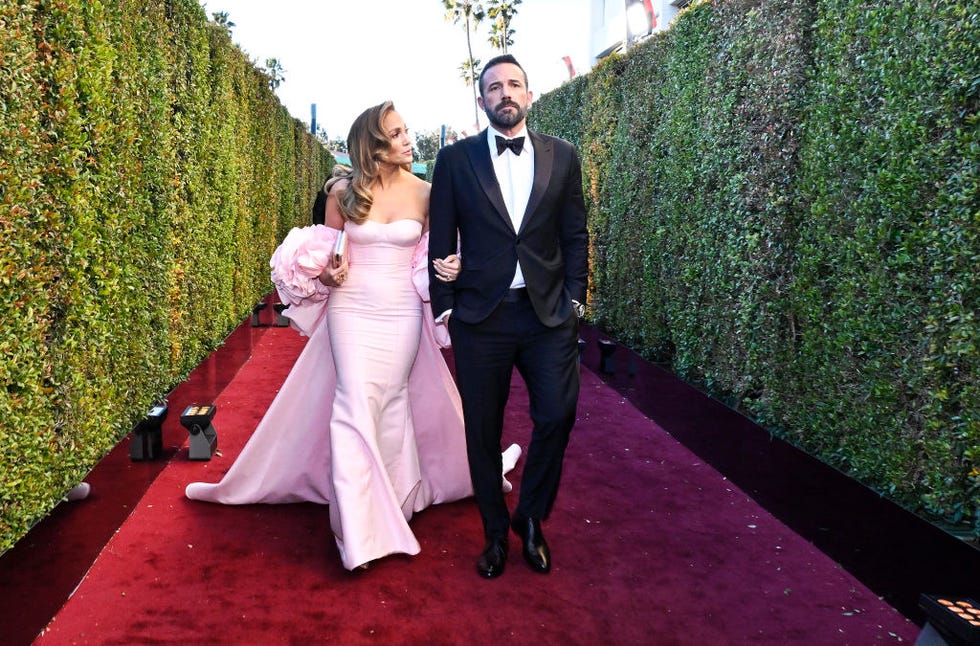 jennifer lopez and ben affleck walk down a red carpet between two green hedges, she wears a pink strapless gown with a long off the shoulder coat, he wears a black tuxedo