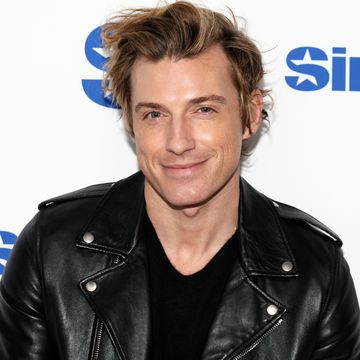 jeremiah brent, a man stands smiling at the camera, he has coiffed blonde hair and wears a black tshirt with black leather jacket