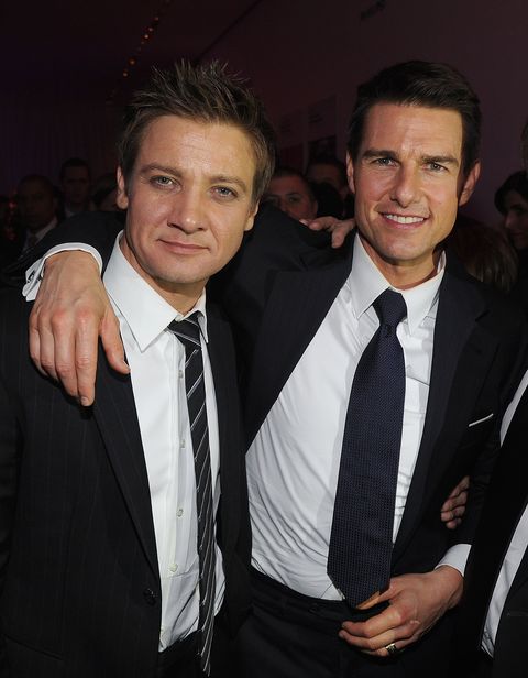 jeremy renner and tom cruise pose for a photo, both smile at the camera and are wearing dark suits with ties, cruise has his arm around renner