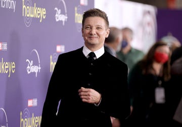jeremy renner smiles past the camera on a red carpet, he is wearing a black jacket, white collared shirt, and black tie, to his left is a disney plus backdrop and behind him are blurry people