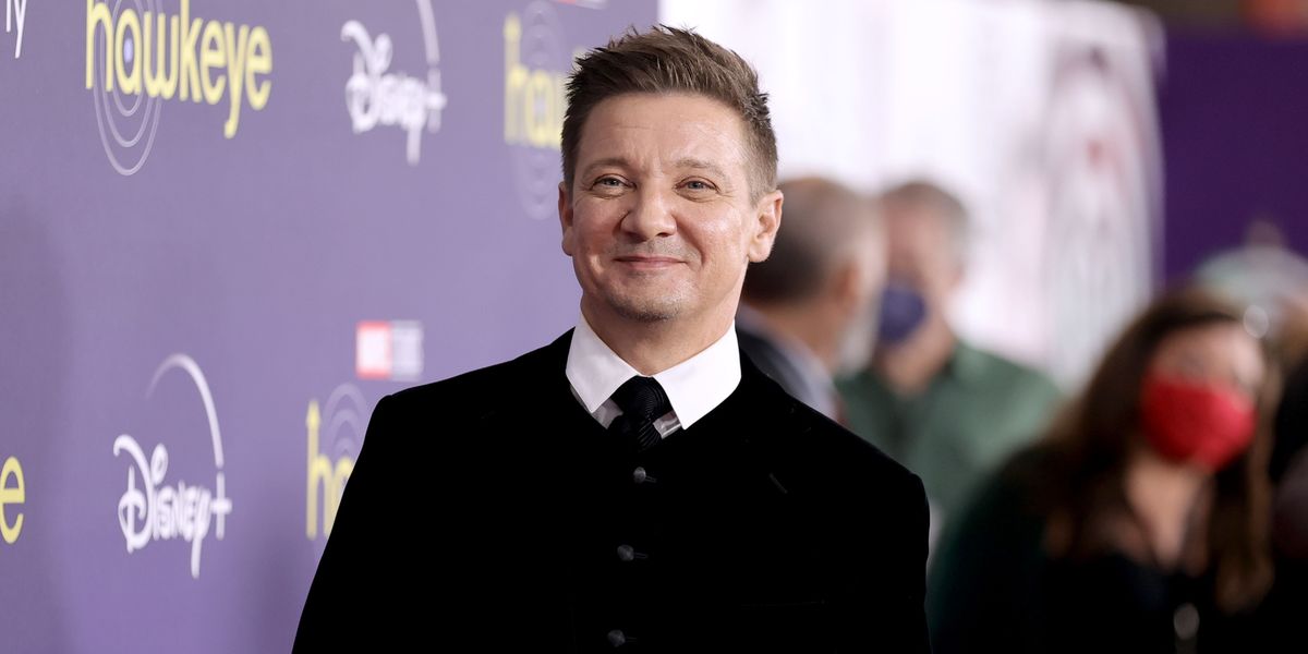 jeremy renner smiles past the camera on a red carpet, he is wearing a black jacket, white collared shirt, and black tie, to his left is a disney plus backdrop and behind him are blurry people