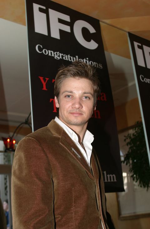 jeremy renner looks at the camera with a neutral expression and stands in front of a banner, he is wearing a brown suit jacket and white collared shirt unbuttoned at the neck