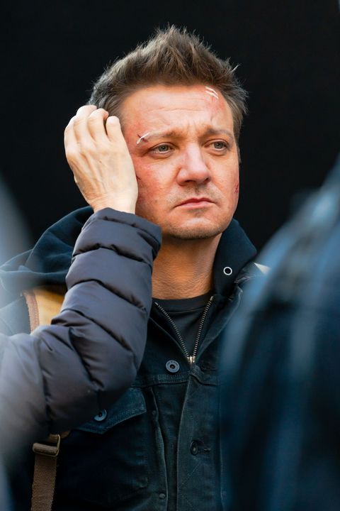 jeremy renner gets a makeup touchup on set, he looks off camera to the right as someone off camera fixes his hair, his face has fake bandages and scrapes