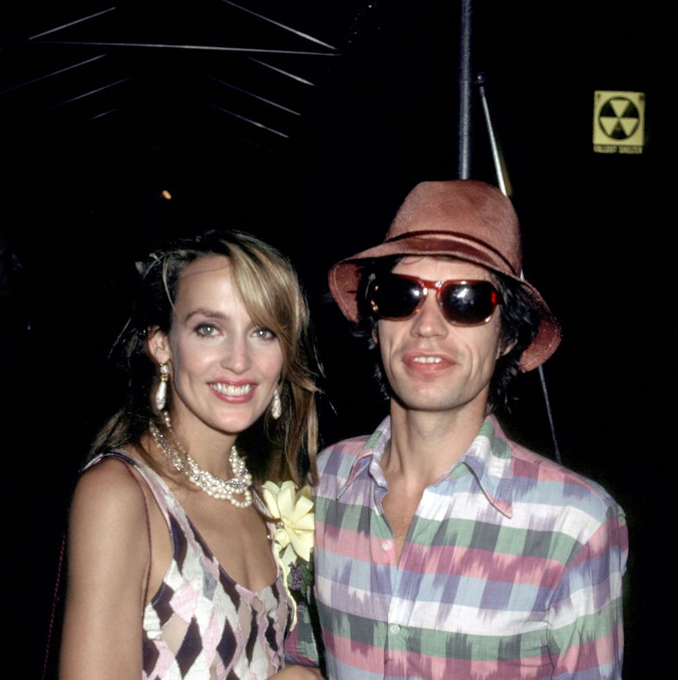 jerry hall and mick jagger smile at the camera while standing together, she wears a chevron patterned sleeveless dress and pearl jewelry, he wears a multicolor striped collared shirt, sunglasses and a dusty red hat