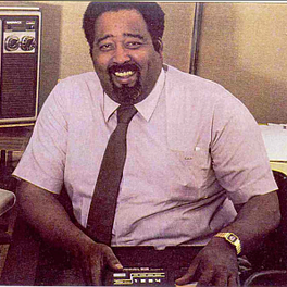 jerry lawson sits at a desk in an office and holds a device on his lap, he is wearing a mauve short sleeve collared shirt and brown tie with a gold watch