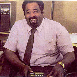 jerry lawson sits at a desk in an office and holds a device on his lap, he is wearing a mauve short sleeve collared shirt and brown tie with a gold watch