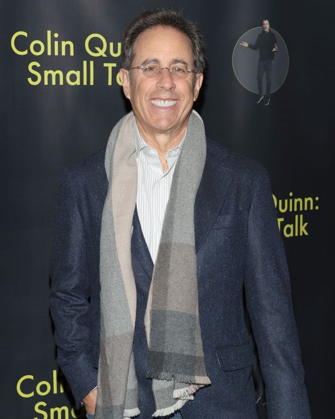 jerry seinfeld smiling for a photo at a premiere event wearing a suit jacket and scarf
