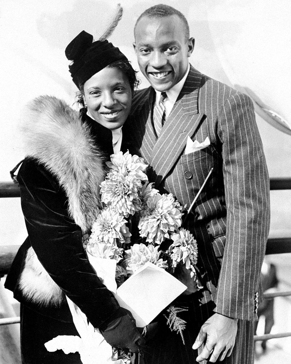 ruth owens and jesse owens stand together and smile for photos, she holds a large bouquet of flowers and wears a dark hat, matching coat and fur stole, he wears a pinstriped suit and tie