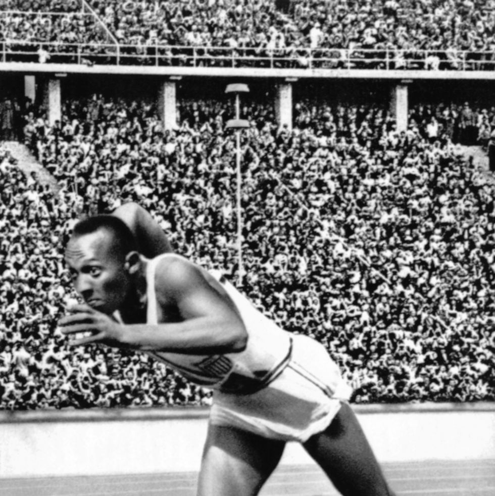 jesse owens leaps from the starting line on a track with a crowd watching from the grandstand, he wears a sleeveless tank top and shorts