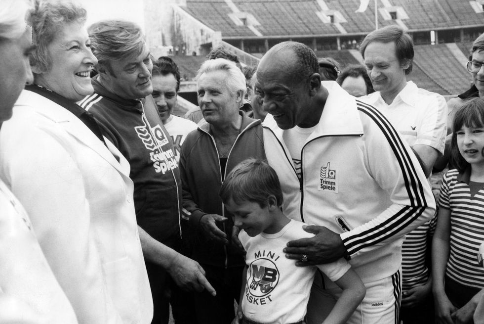 jesse owens smiles as he holds the shoulders of a boy standing in front of him, the two stand in the center of a crowd of people at a stadium
