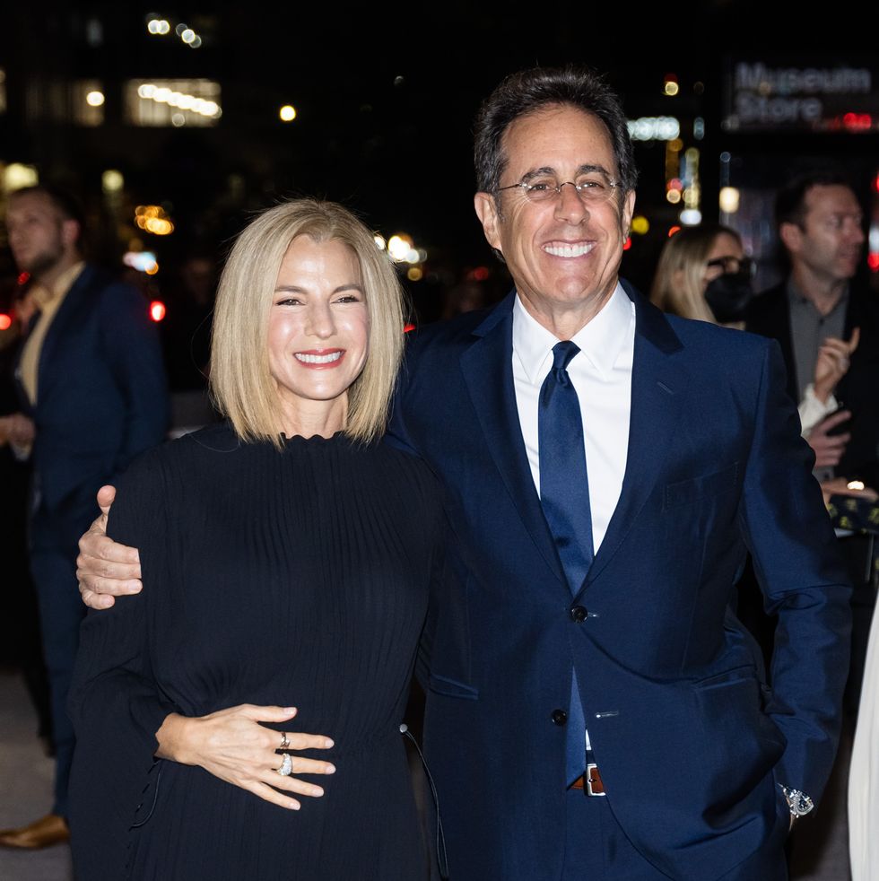 jessica seinfeld and jerry seinfeld smile at a camera on a sidewalk, she wears a long dark dress and he wears a royal blue suit with white collared shirt and blue tie, he has his arm around her