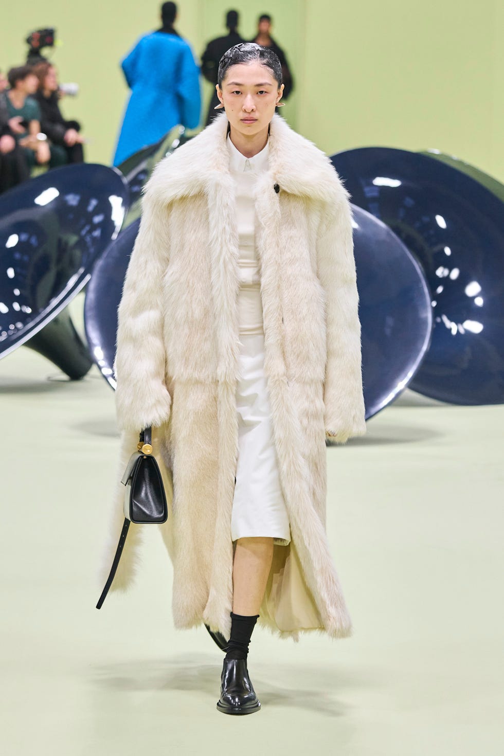 Fashion model on runway wearing luxurious fur coat and holding a small bag