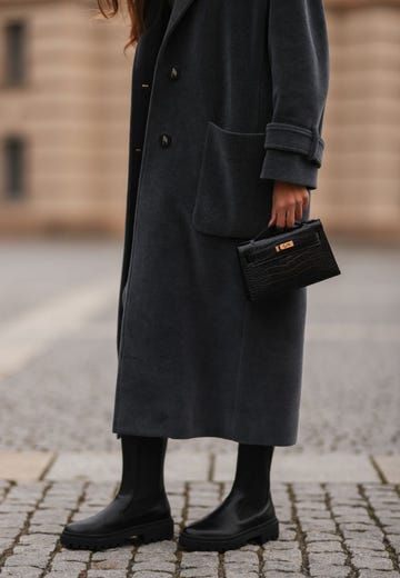 a woman wearing black lug sole chelsea boots with a long grey coat while holding a small black croc top handle bag on a cobblestone street in berlin in a roundup of the most comfortable chelsea boots 2024