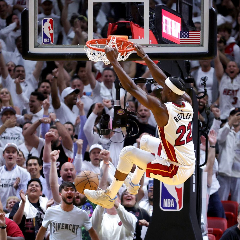jimmy butler, wearing a white miami heat jersey, dunks a basketball into the net at a basketball court, as many fans in white shirts cheer him on in the background