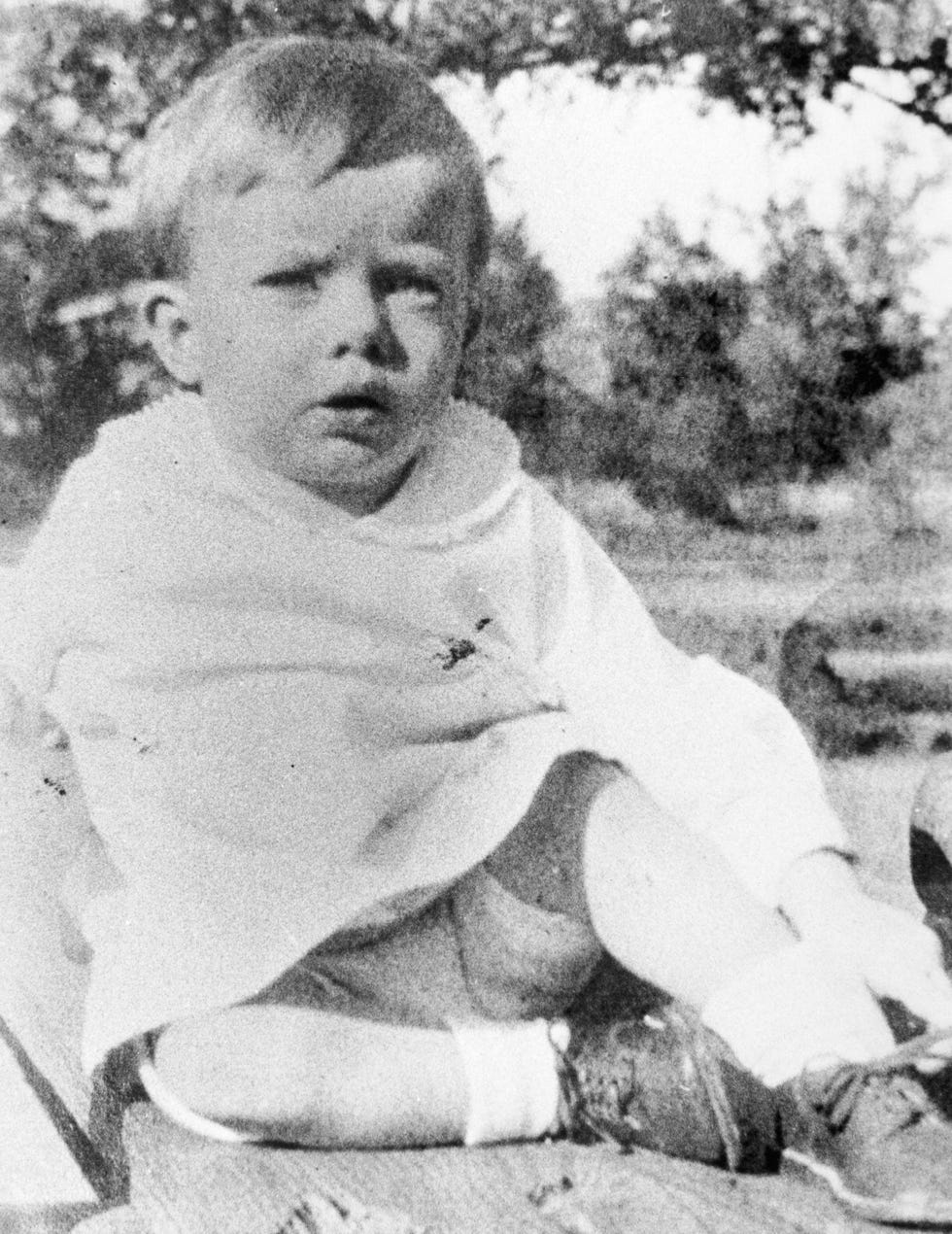 jimmy carter as a young child, the photo is black and white, he sits on what looks like a picnic blanket wearing a light colored outfit, white socks that rise to his mid calf and leather looking shoes with laces, his right leg is crossed in front of his body and his left leg is bent with his knee in the air, he has a confused look on his face