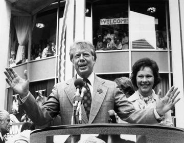 jimmy carter wearing a suit and tie and speaking into a microphone at a podium, standing next to his wife rosalynn, with supporters in stands behind him