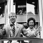 jimmy carter wearing a suit and tie and speaking into a microphone at a podium, standing next to his wife rosalynn, with supporters in stands behind him