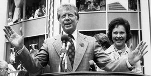 jimmy carter wearing a suit and tie and speaking into a microphone at a podium, standing next to his wife rosalynn, with supporters in stands behind him