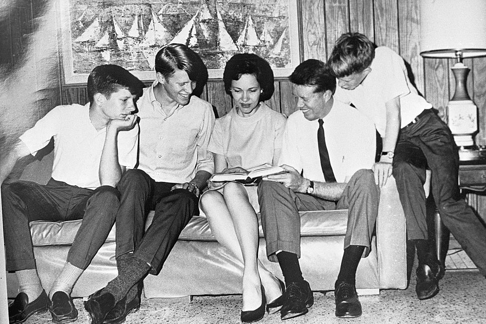chip, jack, rosalynn, jimmy, and jeff carter sit on and around a couch and look at a book that rosalynn holds