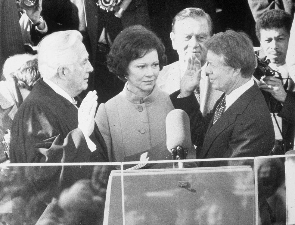 rosalynn carter stands and looks at jimmy carter whose right hand is raised as he faces earl burger whose right hand is also raised, in front of them is a podium with a large microphone, and several people are behind them