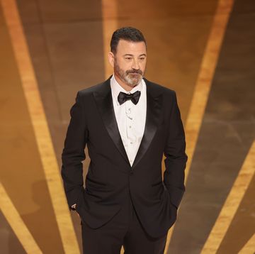 jimmy kimmel stands on a stand with his hands in his pockets, he wears a black tuxedo with a bowtie and a white collard shirt
