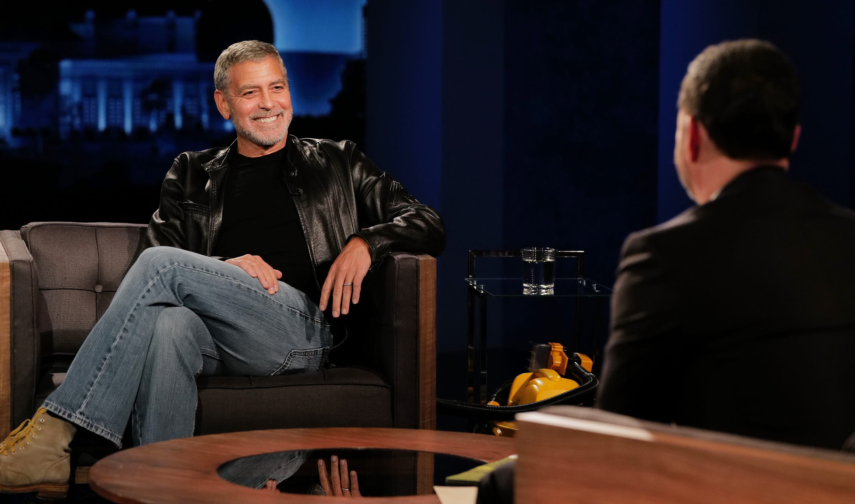 george clooney smiles as he sits in an armchair with his legs crossed, he wears a black leather jacket, black shirt and jeans