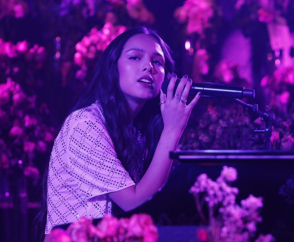 olivia rodrigo sits at a piano and sings into a microphone, she wears several rings and a bejeweled shirt