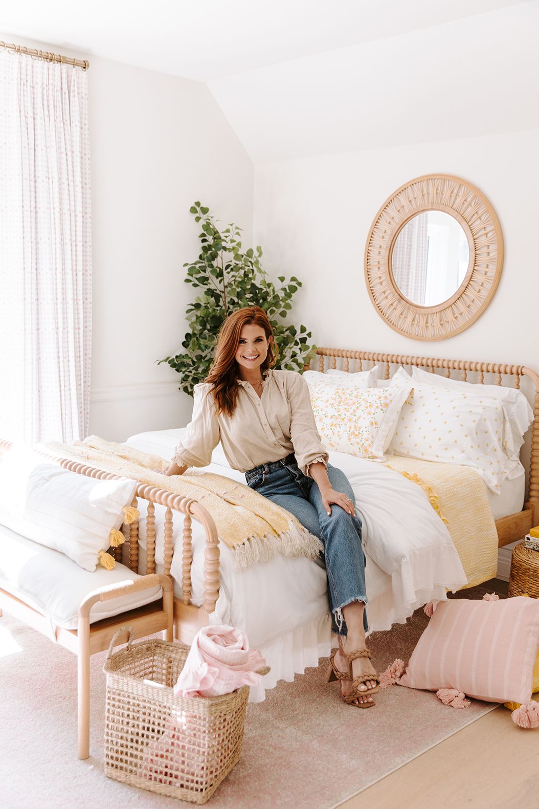 joanna garcia swisher sitting on spindle frame bed with yellow and white linens with basket holding pink blanket and pink fringe pillow at her feet