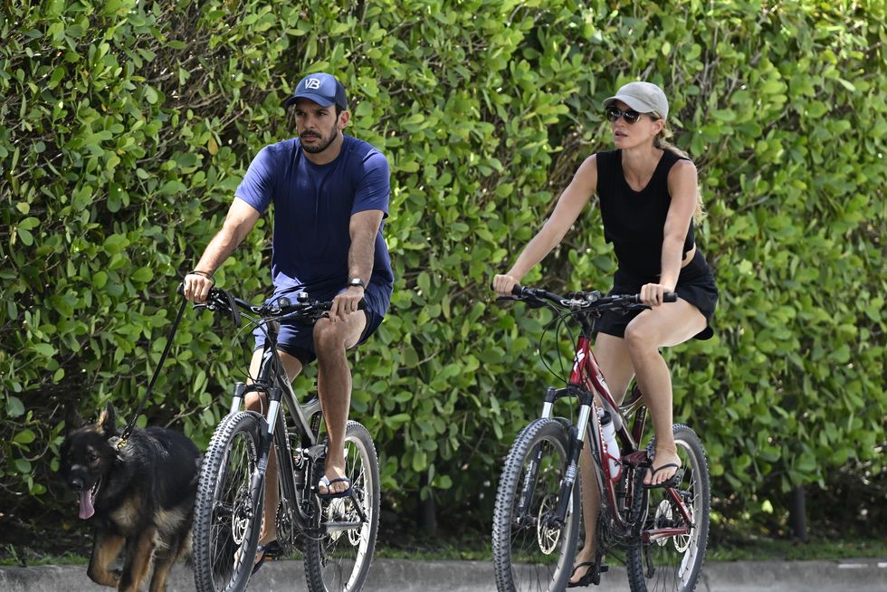 joaquim valente and gisele bundchen ride bikes outside on a street, he holds the leash of a dog walking alongside