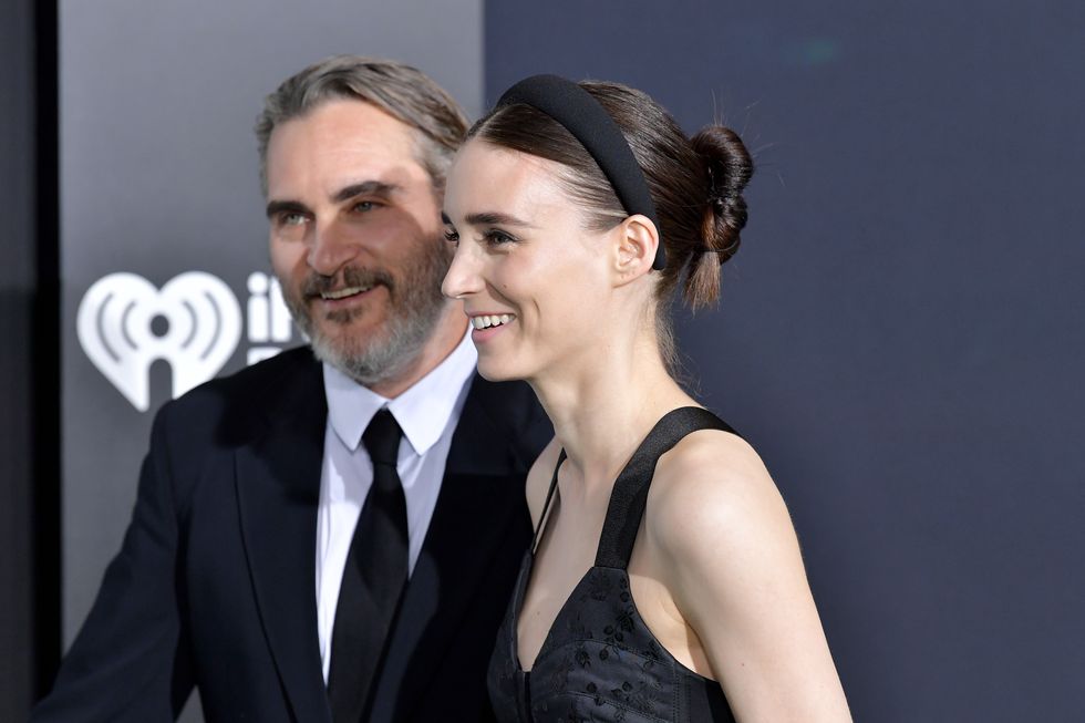 joaquin phoenix, wearing a black suit and tie, and rooney mara, wearing a black dress, smile and look off camera