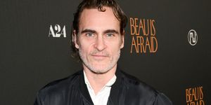 joaquin phoenix smiles at the camera, he wears a black jacket and white collared shirt, his hands are in his jacket pockets and he stands in front of a black background